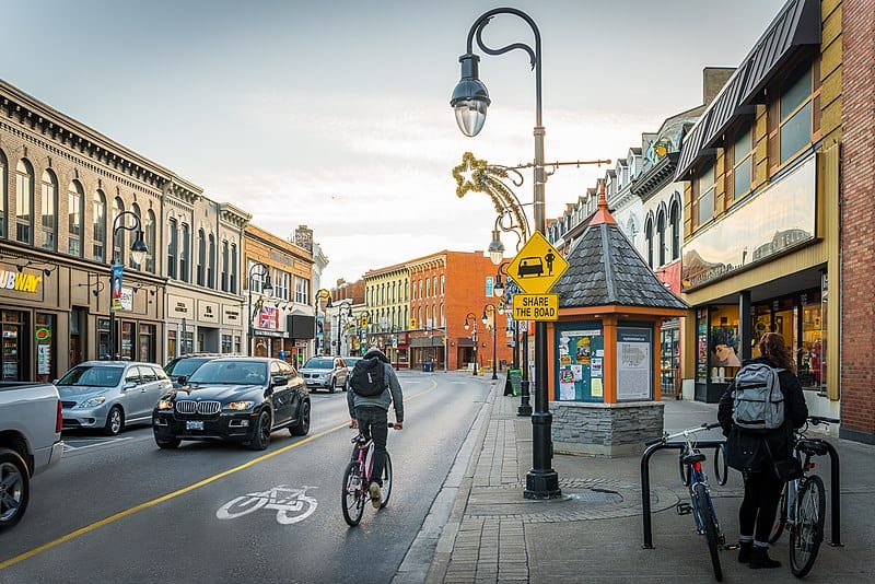 A roadway framed by brick two-storey commercial buildings. The road has two lanes with cars and a contra-flow bikelane with a cyclist in it.