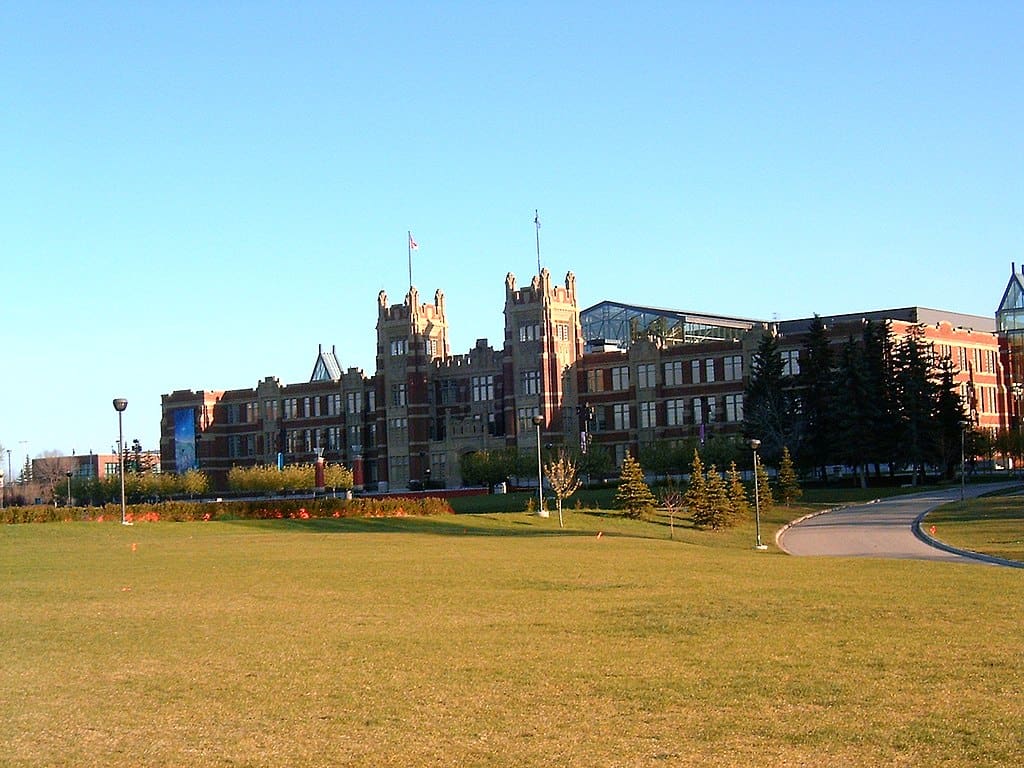 A classic brick building with twin towers at its centre rests at the back of a large lawn, under a blue sky. (SAIT Campus, Alberta)