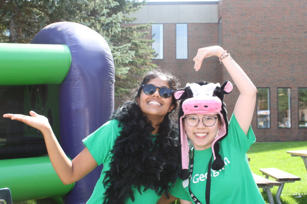Two female Georgian College students pose for a photo during a school event. There is a bouncy castle behind them, on the campus lawn.