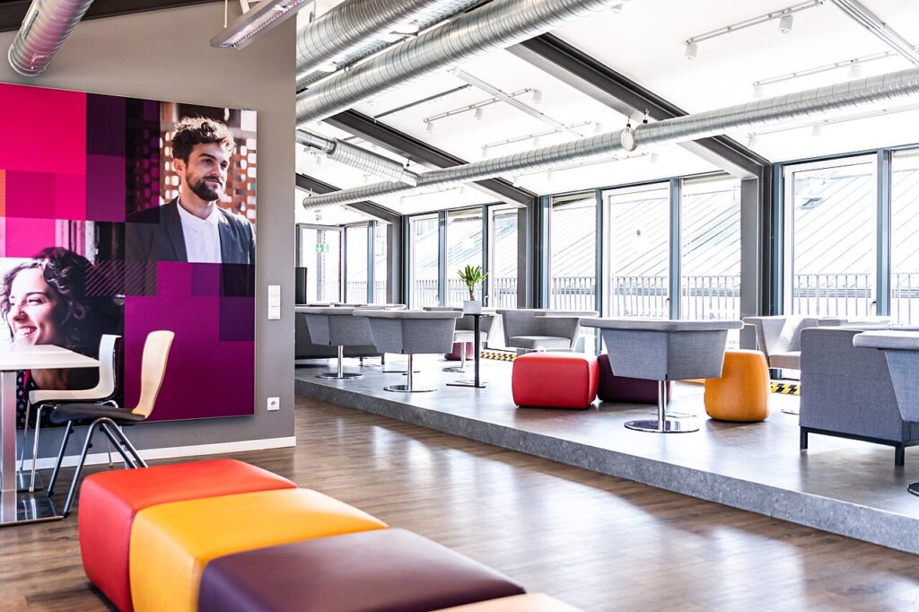 The interior of a Macromedia University building in Munich: glass windows and a partial roof of glass make the space bright. It's filled with tables and bench/pouf seats in greys and bright colours.