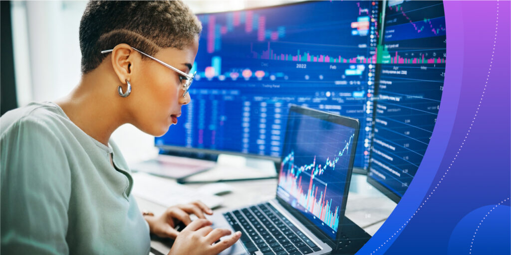 A young woman works at a laptop on a table, which also holds brightly-lit screens with data and projections.