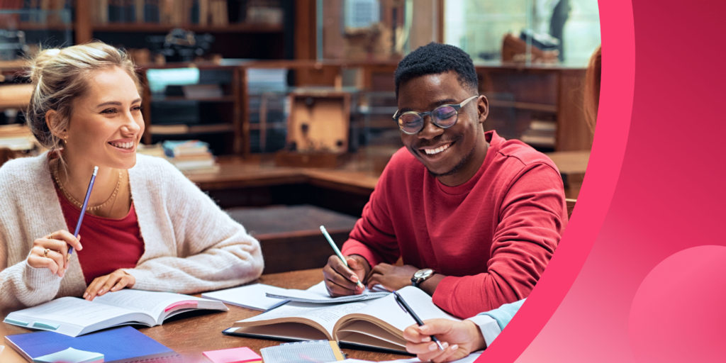 Two students sit at a table in a library, studying together and smiling with their books open