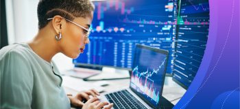 A young woman works at a laptop on a table, which also holds brightly-lit screens with data and projections.