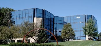 A multistorey glass building rests on a green hill, surrounded by trees and a wheel sculpture.