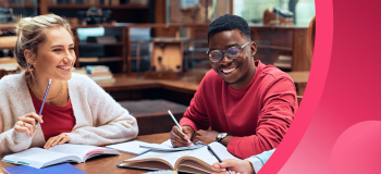 Two students sit at a table in a library, studying together and smiling with their books open