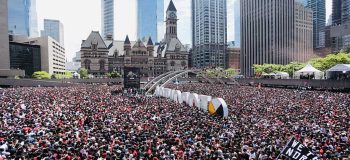 Toronto's downtown on We The North Day after the Raptors' win in 2019: tall steel and glass towers frame a sea of people standing in the public square in front of City Hall. Most are in red and black clothing.