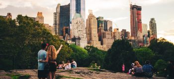 A view of Manhattan's downtown (all the skyscrapers, grey cloudy sky) from the rock formation in Central Park. A young couple with their backs to the camera point at the skyscrapers.